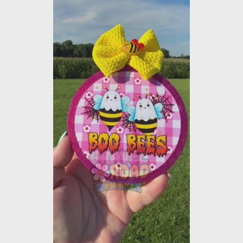 A hand holds a circular Halloween-themed accessory featuring two cartoon bees on a pink gingham background with the words 'BOO BEES' and a large yellow knitted bow on top. The background is an outdoor grassy lawn.