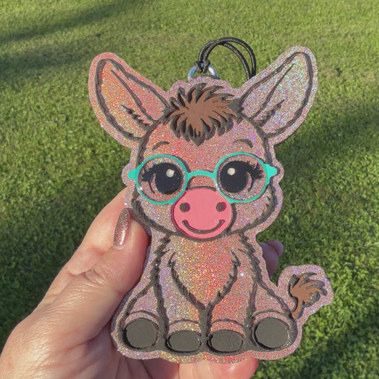 A video shows a hand holding a glittery donkey-shaped car freshie outdoors on a green grass background. The donkey has turquoise glasses, a pink snout, and black hooves.