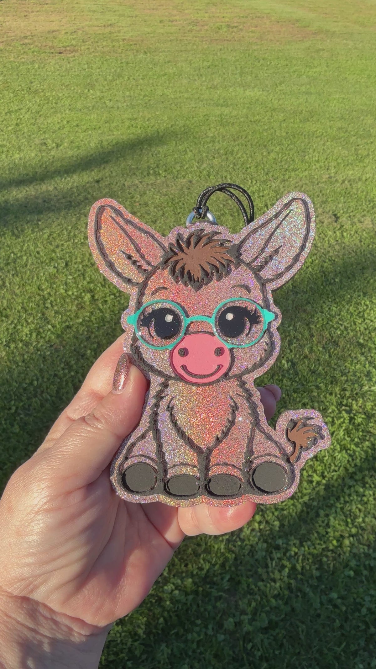 A video shows a hand holding a glittery donkey-shaped car freshie outdoors on a green grass background. The donkey has turquoise glasses, a pink snout, and black hooves.