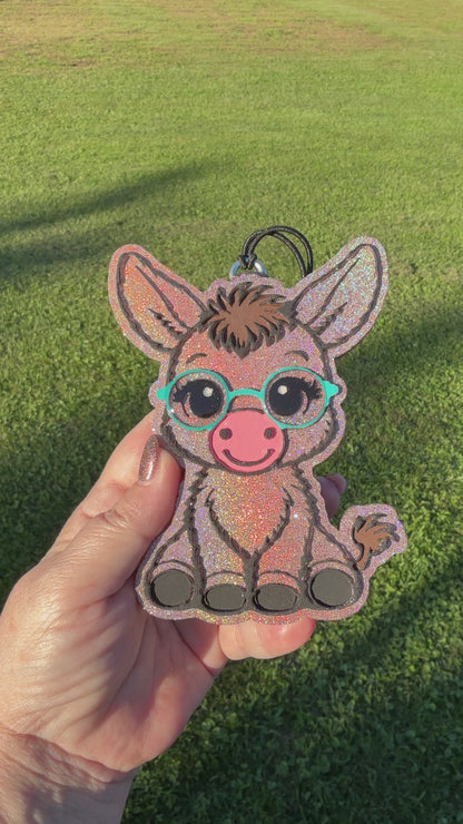 A video shows a hand holding a glittery donkey-shaped car freshie outdoors on a green grass background. The donkey has turquoise glasses, a pink snout, and black hooves.