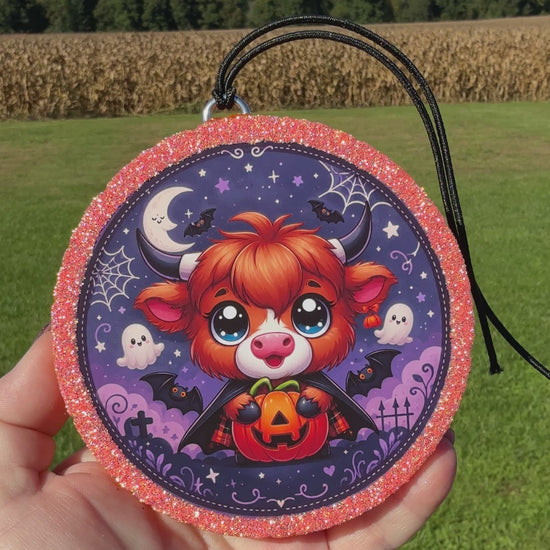 A video showing a Cute Halloween Highland cow freshie with a glittery pink border, black hanging cord, and a spooky purple background featuring stars, bats, ghosts, spider webs, and gravestones.