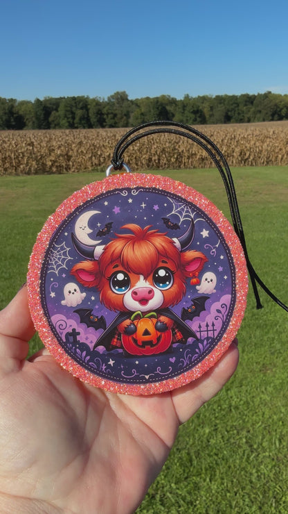 A video showing a Cute Halloween Highland cow freshie with a glittery pink border, black hanging cord, and a spooky purple background featuring stars, bats, ghosts, spider webs, and gravestones.