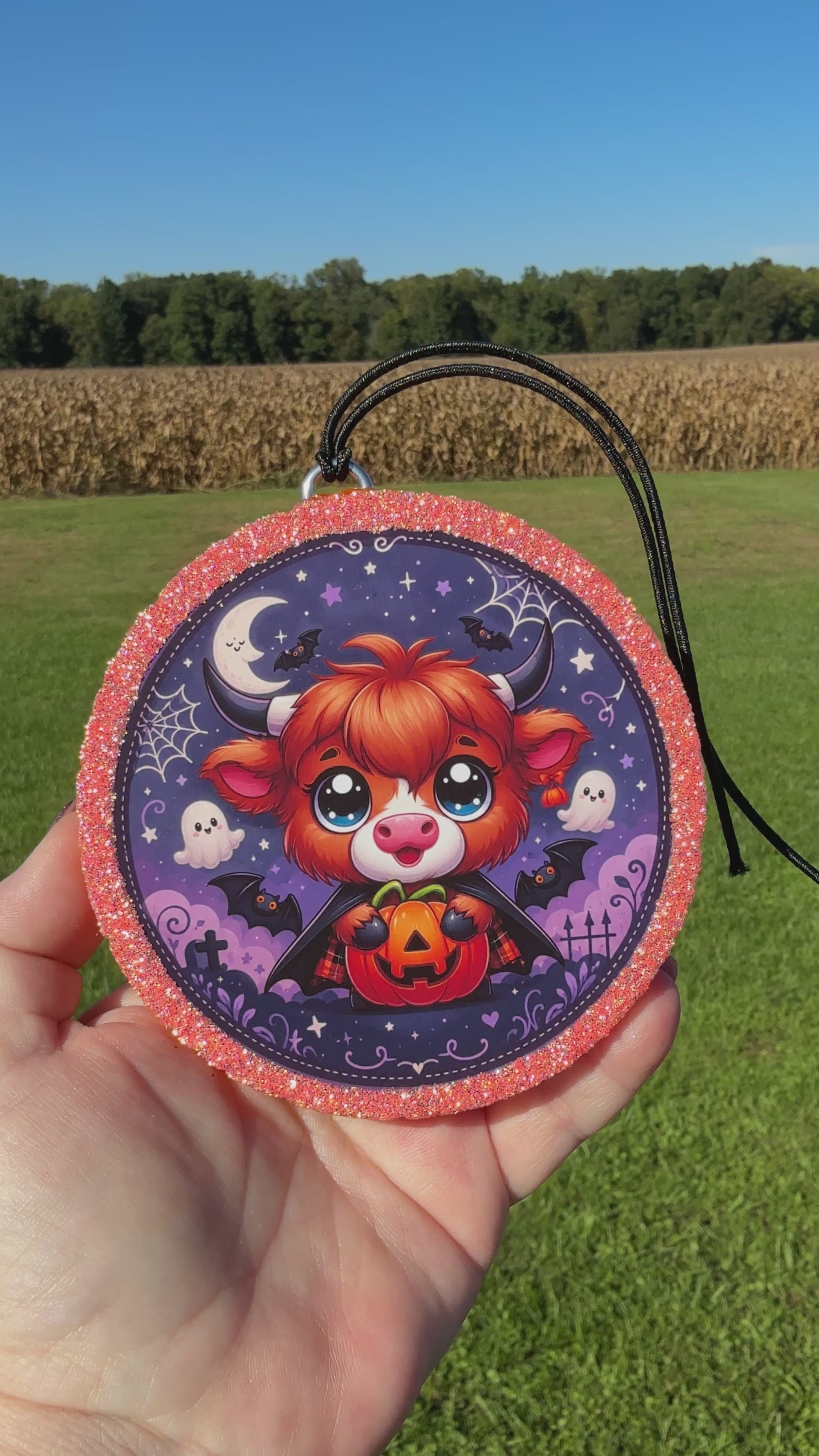 A video showing a Cute Halloween Highland cow freshie with a glittery pink border, black hanging cord, and a spooky purple background featuring stars, bats, ghosts, spider webs, and gravestones.