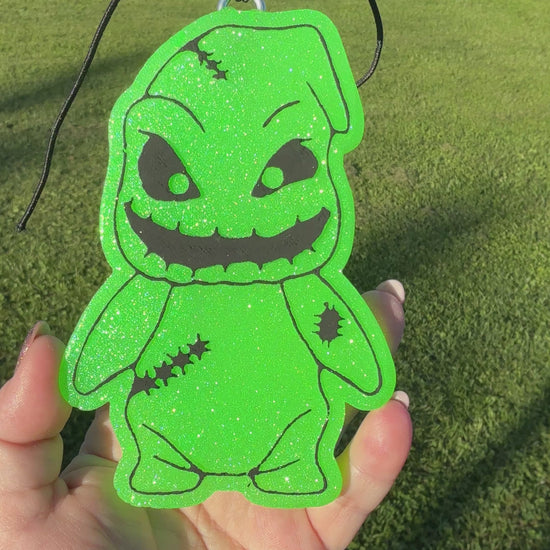 A video shows a hand holding a bright green glittery character-shaped air freshener or car freshie with black stitched details and a black hanging cord, set against an outdoor grassy field background.