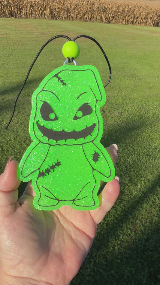 A video shows a hand holding a bright green glittery character-shaped air freshener or car freshie with black stitched details and a black hanging cord, set against an outdoor grassy field background.