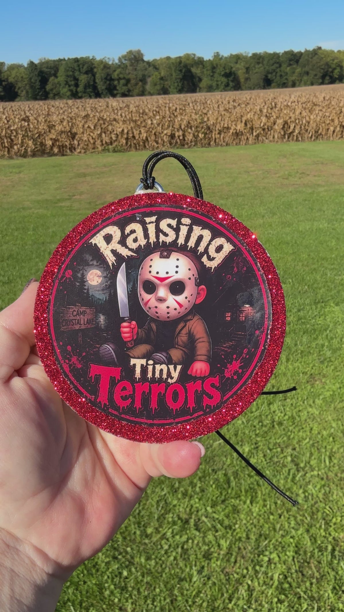 A video showing a Cartoon child horror‑themed freshie with a glittery red border, black hanging cord, and “Raising Tiny Terrors” text in front of a moonlit forest and camp sign