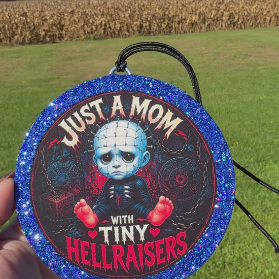 A video showing a Cartoon baby horror‑themed freshie with a glittery blue border, black hanging cord, and “Just a Mom With Tiny Hellraisers” text on a dark chain‑themed background.