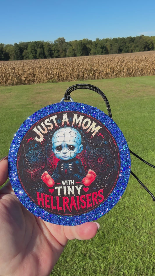 A video showing a Cartoon baby horror‑themed freshie with a glittery blue border, black hanging cord, and “Just a Mom With Tiny Hellraisers” text on a dark chain‑themed background.